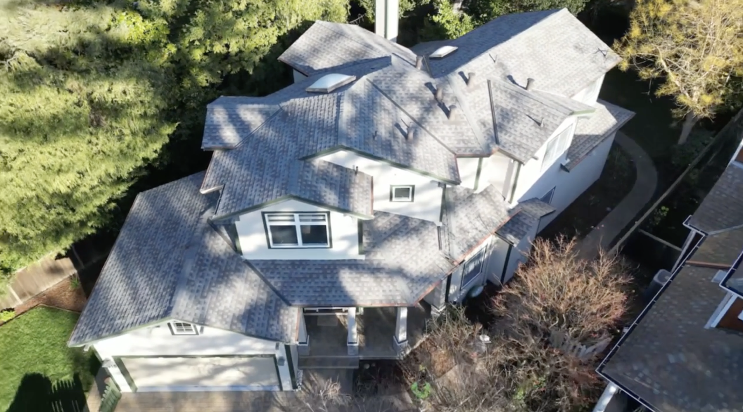 Aerial view of a large, two-story house with a complex, gray-shingled roof. The house is surrounded by tall trees and has a curved pathway leading to the entrance. It's situated in a residential area with neighboring homes.